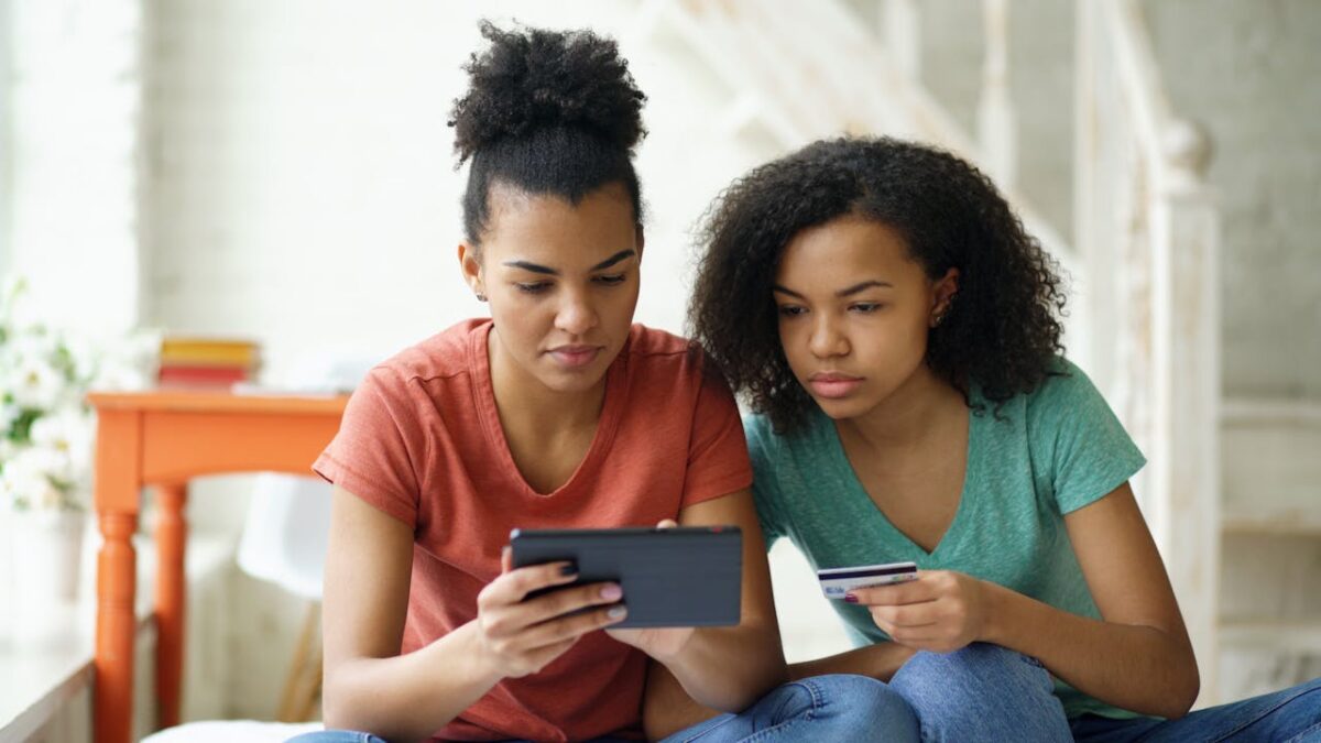 Two young women using a tablet and credit card for online shopping in a cozy indoor setting.