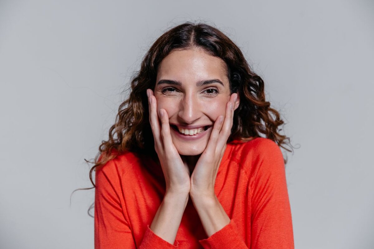 Happy woman in a vibrant orange sweater smiling with hands on her face.